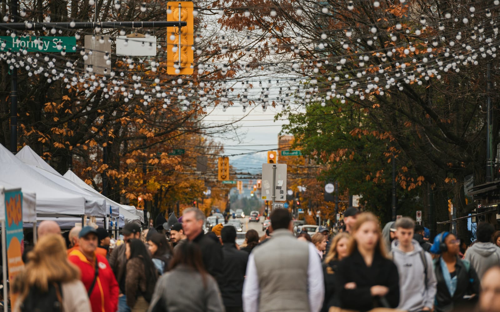 Visitors exploring an outdoor street market beneath string lights and autumn trees in downtown Vancouver.
